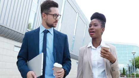 African-american-businesswoman-and-caucasian-businessman-holding-coffee-cups-and-talking-while-walking-down-the-street