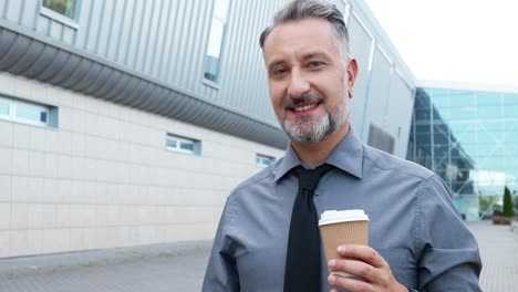 Close-up-view-of-gray-haired-businessman-holding-coffee-cup-and-looking-at-camera-in-the-street