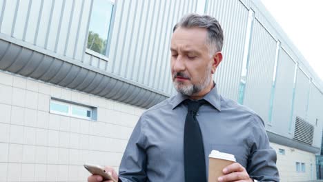Close-up-view-of-gray-haired-businessman-holding-coffee-cup-and-using-smartphone-in-the-street