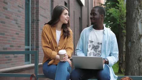 Camera-zooming-on-african-american-man-and-caucasian-woman-using-laptop-and-holding-coffe-cup-sitting-in-the-street-near-the-college