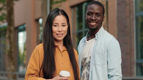 African-american-and-asian-students-looking-at-the-camera-while-standing-near-the-university-in-a-break