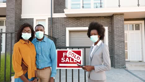 African-American-young-happy-couple-in-medical-masks-standing-outdoor-at-new-house-with-female-real-estate-agent-looking-at-the-camera
