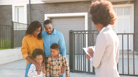 Young-African-American-family-with-two-small-kids-buying-house-at-suburbs-and-talking-with-female-real-estate-agent-outdoors