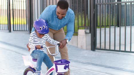 African-American-father-teaching-small-girl-in-helmet-riding-on-bike-at-street-in-suburb