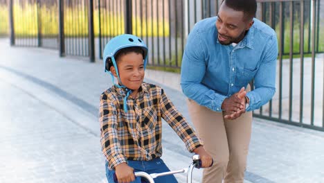 Padre-Afroamericano-Enseñando-A-Un-Niño-Pequeño-Con-Casco-Montando-En-Bicicleta-En-La-Calle-En-El-Suburbio
