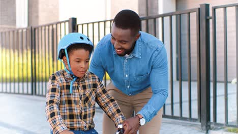 Happy-African-American-father-teaching-small-boy-in-helmet-riding-on-bike-at-street-in-outskirt