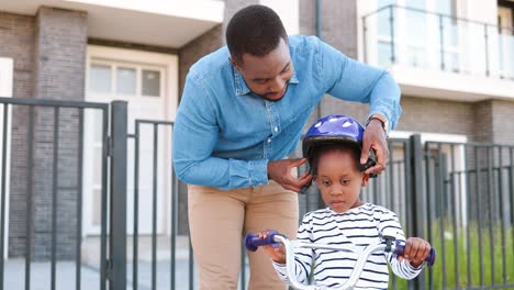 Afroamerikanischer-Vater,-Der-Einem-Kleinen-Hübschen-Mädchen-Einen-Helm-Aufsetzt-Und-Das-Fahrradfahren-Auf-Der-Straße-Lehrt