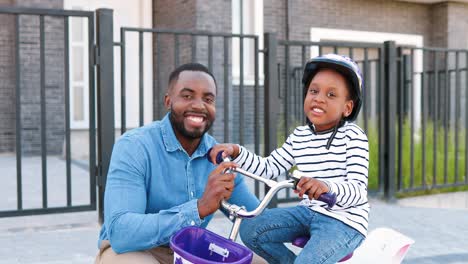Portrait-of-African-American-small-girl-happy-daughter-in-helmet-sitting-on-bike-with-young-father-smiling-to-camera
