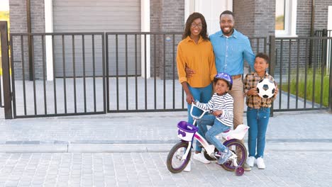 Retrato-De-Una-Feliz-Familia-Afroamericana-Con-Niños-Pequeños-Parados-En-La-Calle-Cerca-De-La-Casa-En-El-Suburbio-Y-Sonriendo-A-La-Cámara