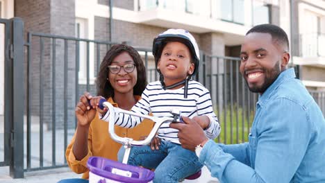 Portrait-shot-of-young-African-American-happy-family-with-small-cute-girl-on-bike-smiling-to-camera-outdoor