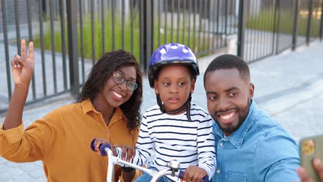 Portrait-of-African-American-family-and-cute-little-daughter-in-helmet-sitting-on-bike-smiling-to-smartphone-camera-and-taking-selfie-photos