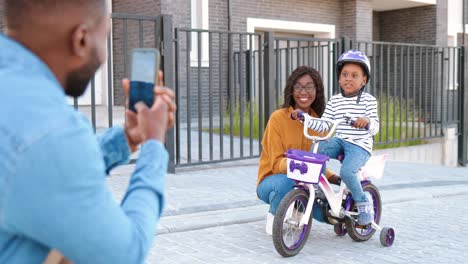 Back-view-on-African-American-father-taking-photo-with-smartphone-camera-of-wife-and-daughter-on-bike