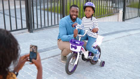 Back-view-on-African-American-mother-taking-photo-with-smartphone-camera-of-husband-and-daughter-on-bike