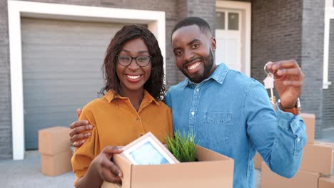 Portrait-of-African-American-young-married-couple-of-man-and-woman-standing-at-new-home,-holding-carton-box-with-stuff-and-showing-key-when-moving-in