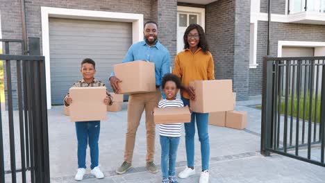 Portrait-of-happy-African-American-family-with-small-children-standing-at-new-house-at-suburb-and-smiling-with-carton-boxes-in-hands