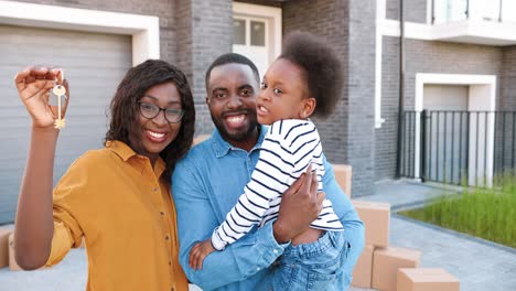 Portrait-of-happy-African-American-family-with-small-cute-girl-on-hands-standing-at-big-house-on-suburb,-smiling-and-showing-key