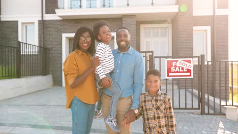 Portrait-of-happy-African-American-family-with-small-children-standing-at-new-house-at-suburb-and-looking-at-camera