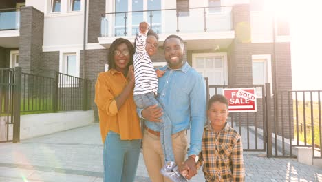 Portrait-of-happy-African-American-family-with-small-children-standing-at-new-house-at-suburb-and-showing-key-to-camera