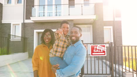 Portrait-of-happy-African-American-family-with-small-child-standing-at-new-house-at-suburb-and-showing-keys-to-camera