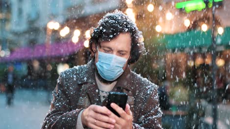 Nahaufnahme-Eines-Kaukasischen-Mannes,-Der-Eine-Gesichtsmaske-Trägt-Und-Ein-Smartphone-Auf-Der-Straße-Benutzt,-Während-Es-In-Weihnachten-Schneit
