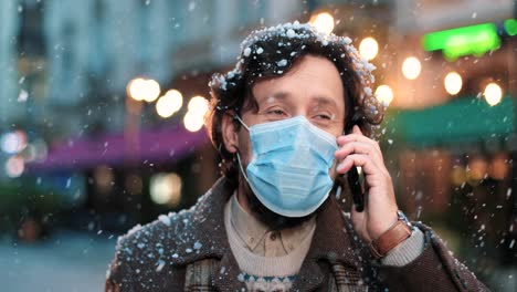 Nahaufnahme-Eines-Kaukasischen-Mannes,-Der-Eine-Gesichtsmaske-Trägt-Und-Auf-Der-Straße-Mit-Dem-Smartphone-Spricht,-Während-Es-In-Weihnachten-Schneit