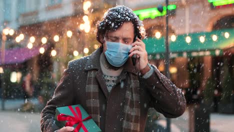 Nahaufnahme-Eines-Kaukasischen-Mannes-Mit-Gesichtsmaske,-Der-Telefoniert-Und-Ein-Geschenk-Auf-Der-Straße-Hält,-Während-Es-Zu-Weihnachten-Schneit