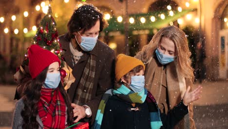 Close-up-view-of-Caucasian-happy-family-wearing-facial-masks-walking-down-the-street-and-holding-Christmas-tree-while-it¬¥s-snowing-in-Christmas
