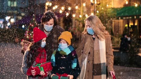 Nahaufnahme-Einer-Kaukasischen-Glücklichen-Familie-Mit-Gesichtsmasken,-Die-Die-Straße-Hinuntergeht-Und-Geschenke-Hält,-Während-Es-Zu-Weihnachten-Schneit