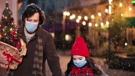 Close-up-view-of-Caucasian-happy-father-and-daughter-wearing-facial-masks-walking-down-the-street-and-holding-Christmas-tree-and-present-while-it¬¥s-snowing-in-Christmas
