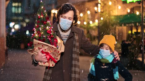 Close-up-view-of-Caucasian-happy-father-and-son-wearing-facial-masks-walking-down-the-street-and-holding-Christmas-tree-and-present-while-it¬¥s-snowing-in-Christmas