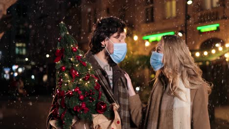 Caucasian-happy-couple-wearing-facial-masks-holding-christmas-tree-and-talking-while-snowing-on-the-street-in-Christmas