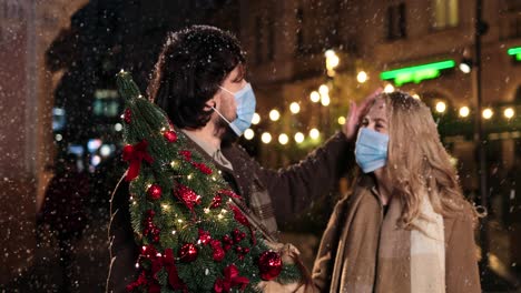 Kaukasisches-Glückliches-Paar,-Das-Gesichtsmasken-Trägt,-Die-Weihnachtsbaum-Halten-Und-Lachen,-Während-Es-Zu-Weihnachten-Auf-Der-Straße-Schneit