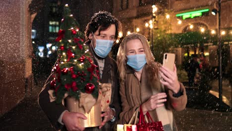 Kaukasisches-Glückliches-Paar,-Das-Gesichtsmasken-Trägt,-Die-Weihnachtsbaum-Halten-Und-Lachen,-Während-Sie-Zu-Weihnachten-Ein-Selfie-Mit-Dem-Smartphone-Auf-Der-Straße-Machen