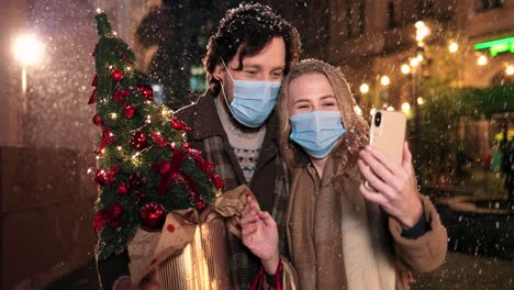 Kaukasisches-Glückliches-Paar,-Das-Gesichtsmasken-Trägt-Und-Weihnachtsbaum-Hält-Und-Winkt,-Während-Es-Zu-Weihnachten-Ein-Video-Mit-Dem-Smartphone-Auf-Der-Straße-Macht
