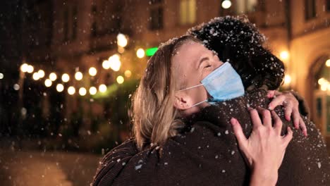 Close-up-view-of-Caucasian-wife-and-husband-wearing-facial-masks-hugging-and-smiling-while-snowing-on-the-street-in-Christmas