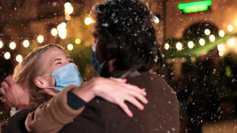 Close-up-view-of-Caucasian-wife-and-husband-wearing-facial-masks-hugging-and-smiling-while-snowing-on-the-street-in-Christmas