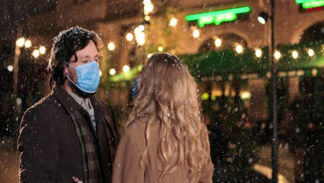Caucasian-happy-couple-wearing-facial-masks-talking-while-snowing-on-the-street-in-Christmas