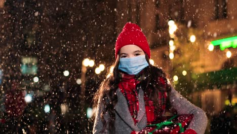 Portrait-of-happy-Caucasian-little-girl-in-scarf-holding-a-present-and-smiling-at-camera-while-it¬¥s-snowing-on-the-street-in-Christmas