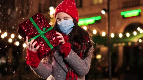 Portrait-of-happy-Caucasian-little-girl-in-scarf-holding-a-present-and-smiling-at-camera-while-it¬¥s-snowing-on-the-street-in-Christmas