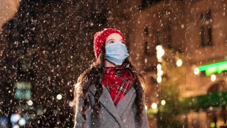 Close-up-view-of-cheerful-little-girl-wearing-facial-mask-standing-on-street-in-winter-and-trying-to-catch-snow-in-Christmas