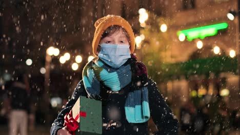 Portrait-of-happy-Caucasian-little-boy-in-scarf-holding-a-present-and-smiling-at-camera-while-it¬¥s-snowing-on-the-street-in-Christmas