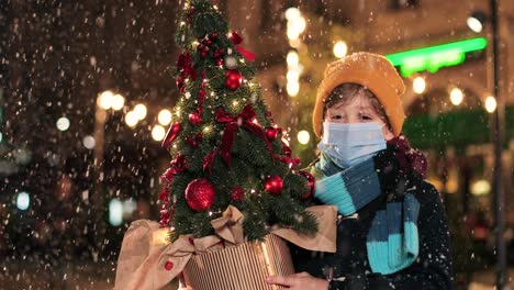 Portrait-of-happy-Caucasian-little-boy-in-scarf-holding-a-Christmas-tree-and-smiling-at-camera-while-it¬¥s-snowing-on-the-street-in-Christmas