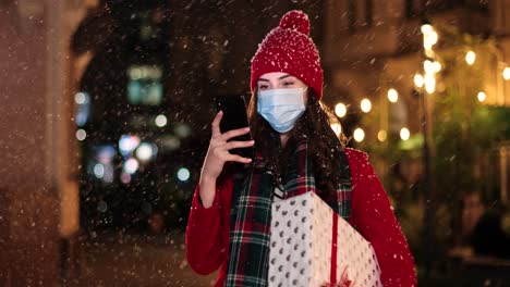 Portrait-of-happy-beautiful-young-woman-typing-on-smartphone-and-holding-a-present-on-the-street-while-it¬¥s-snowing-in-Christmas