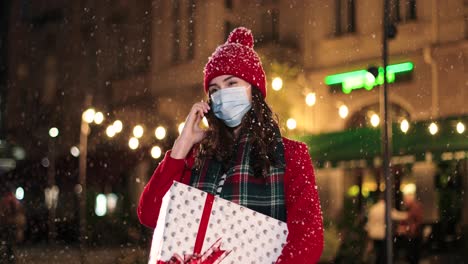 Portrait-of-happy-beautiful-young-woman-talking-on-smartphone-and-holding-a-present-on-the-street-while-it¬¥s-snowing-in-Christmas