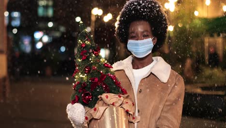 Nahaufnahme-Einer-Fröhlichen-Afroamerikanischen-Frau-Mit-Gesichtsmaske,-Die-Einen-Weihnachtsbaum-Hält-Und-Auf-Der-Straße-In-Die-Kamera-Lächelt,-Während-Es-Zu-Weihnachten-Schneit