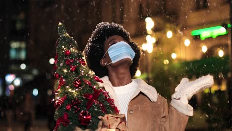 Close-up-view-of-joyful-African-American-woman-wearing-facial-mask-holding-a-Christmas-tree-and-playing-with-the-snow-on-the-street-in-Christmas