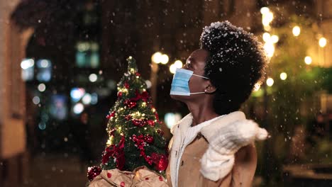 Close-up-view-of-joyful-African-American-woman-wearing-facial-mask-holding-a-Christmas-tree-and-playing-with-the-snow-on-the-street-in-Christmas