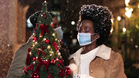 Vista-De-Cerca-De-Una-Alegre-Pareja-Afroamericana-Con-Máscaras-Faciales-Hablando-Y-Sosteniendo-Un-árbol-De-Navidad-Mientras-Nieva-En-La-Calle-En-Navidad
