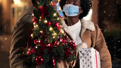Nahaufnahme-Eines-Fröhlichen-Afroamerikanischen-Paares,-Das-Gesichtsmasken-Trägt-Und-Einen-Weihnachtsbaum-Hält,-Während-Es-Zu-Weihnachten-Auf-Der-Straße-Schneit