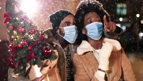 Nahaufnahme-Eines-Fröhlichen-Afroamerikanischen-Paares-Mit-Gesichtsmasken,-Das-Sich-Unterhält-Und-Umschaut,-Während-Es-Zu-Weihnachten-Auf-Der-Straße-Schneit